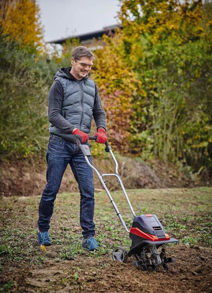 Ein Mann mit Gartenarbeitskleidung schiebt die Akku-Bodenhacke von Einhell durch die Beeterde.